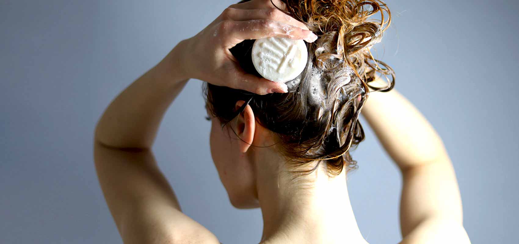Rückansicht einer Frau, die mit einer weißen Shampoobar ihr Haar einschäumt. Die Haare sind nass und schaumig.